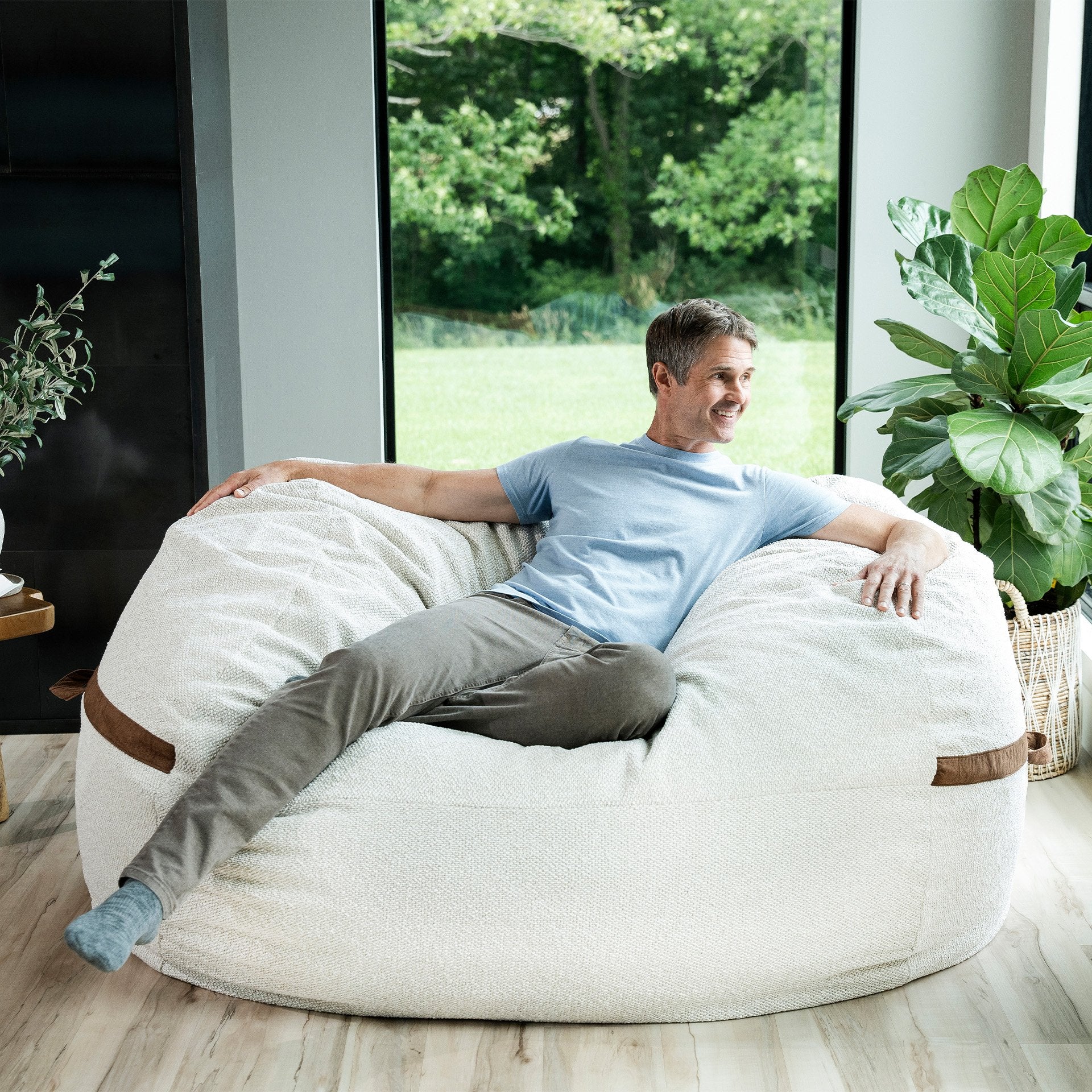 Man sitting on a large white bean bag chair in a modern living room with plants and a window view. #color_sandstone-textured-boucle