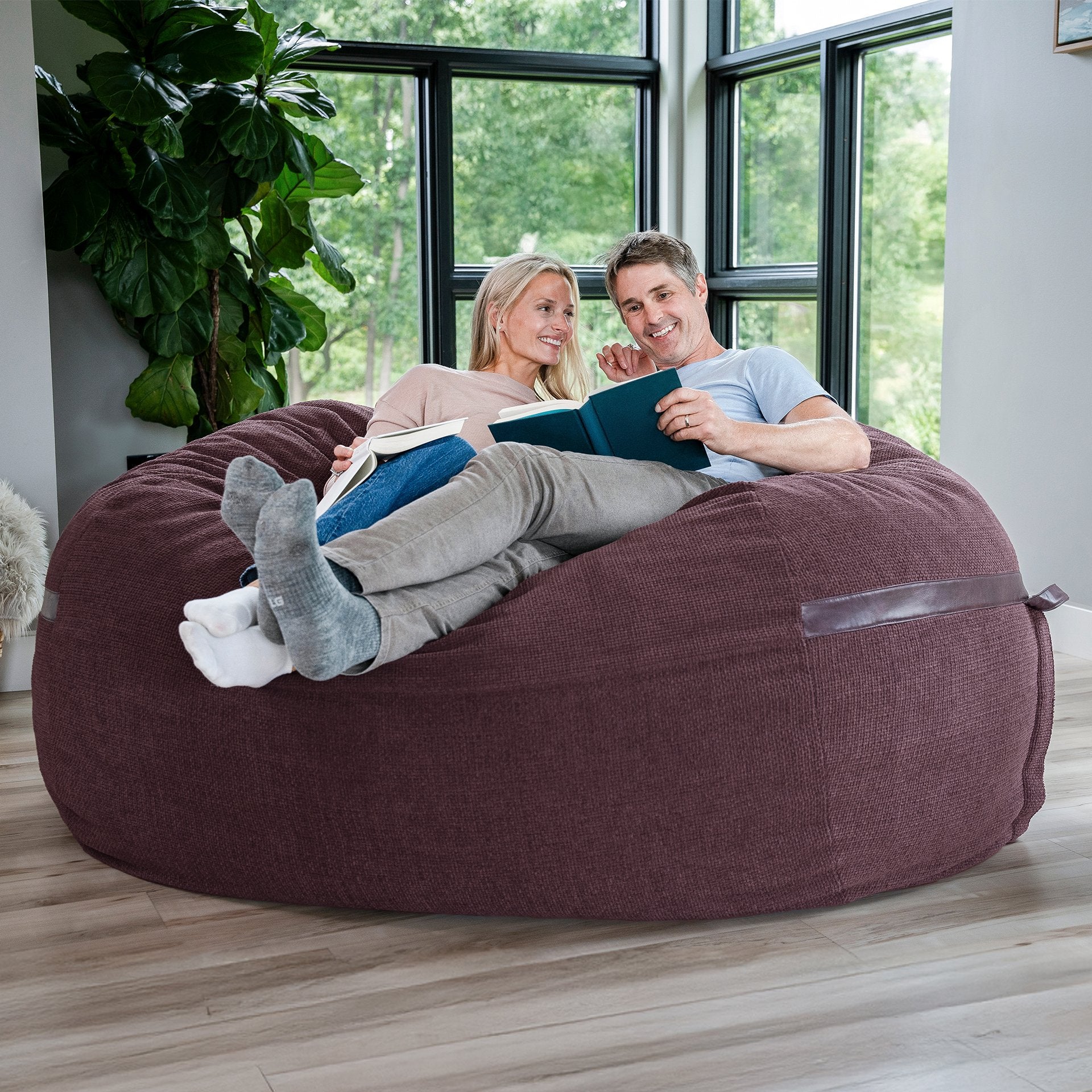 Two people sitting on a large bean bag in a room with large windows and plants. #color_merlot-cloudweave
