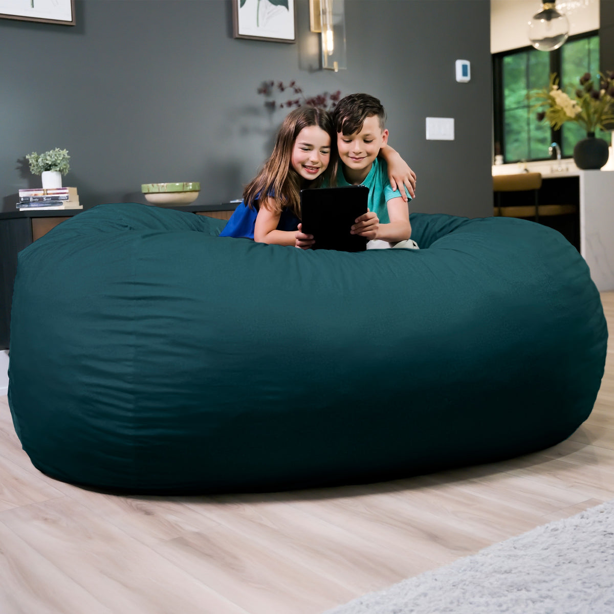 Two children sitting on a large teal bean bag in a living room.