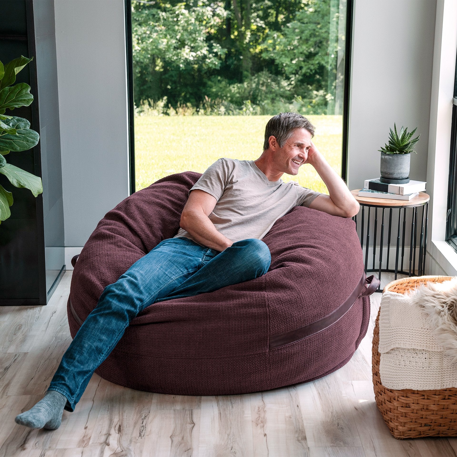 Man sitting on a large brown bean bag chair in a modern living room. #color_merlot-cloudweave