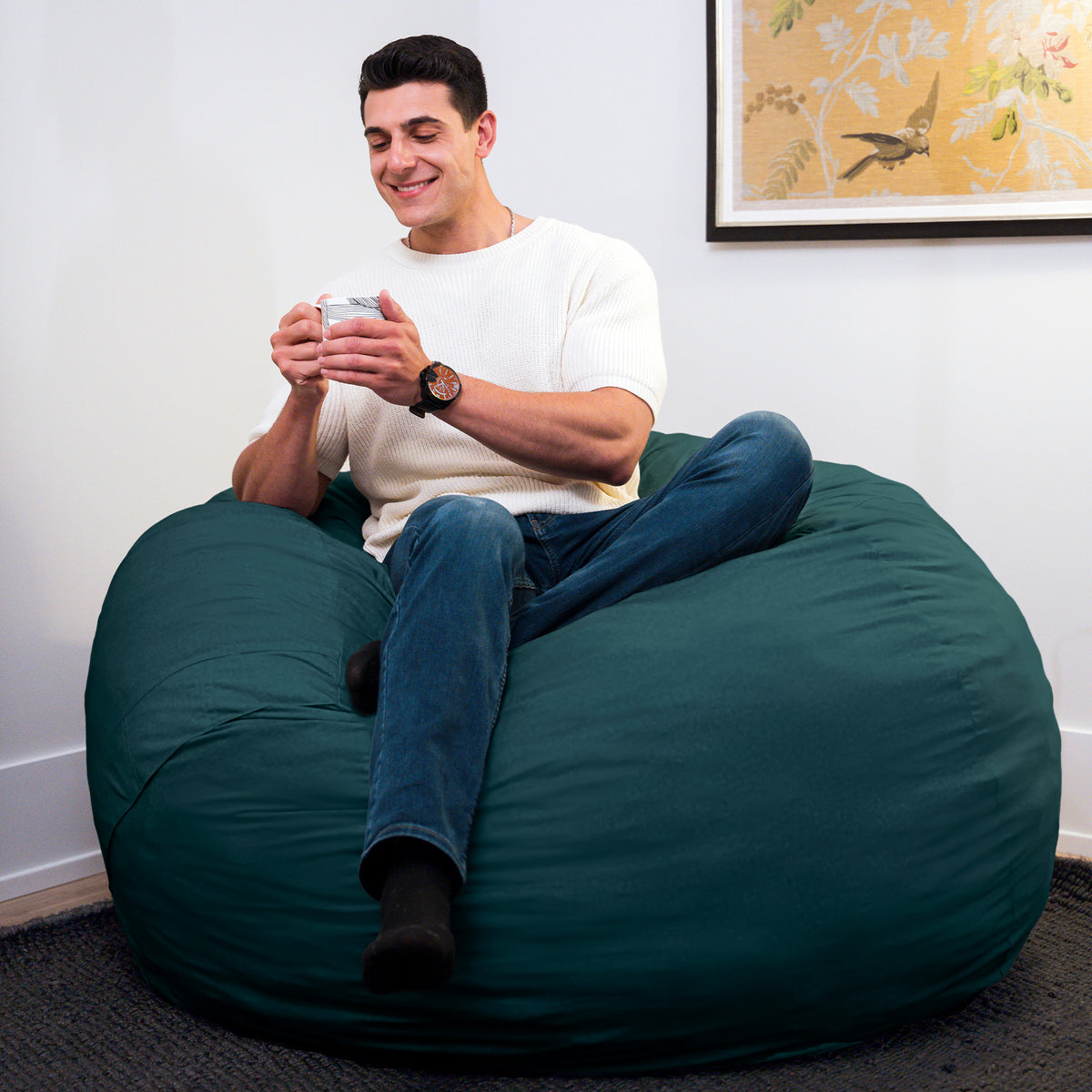 Man sitting on a large green bean bag chair using a smartphone in a room with a framed picture on the wall.