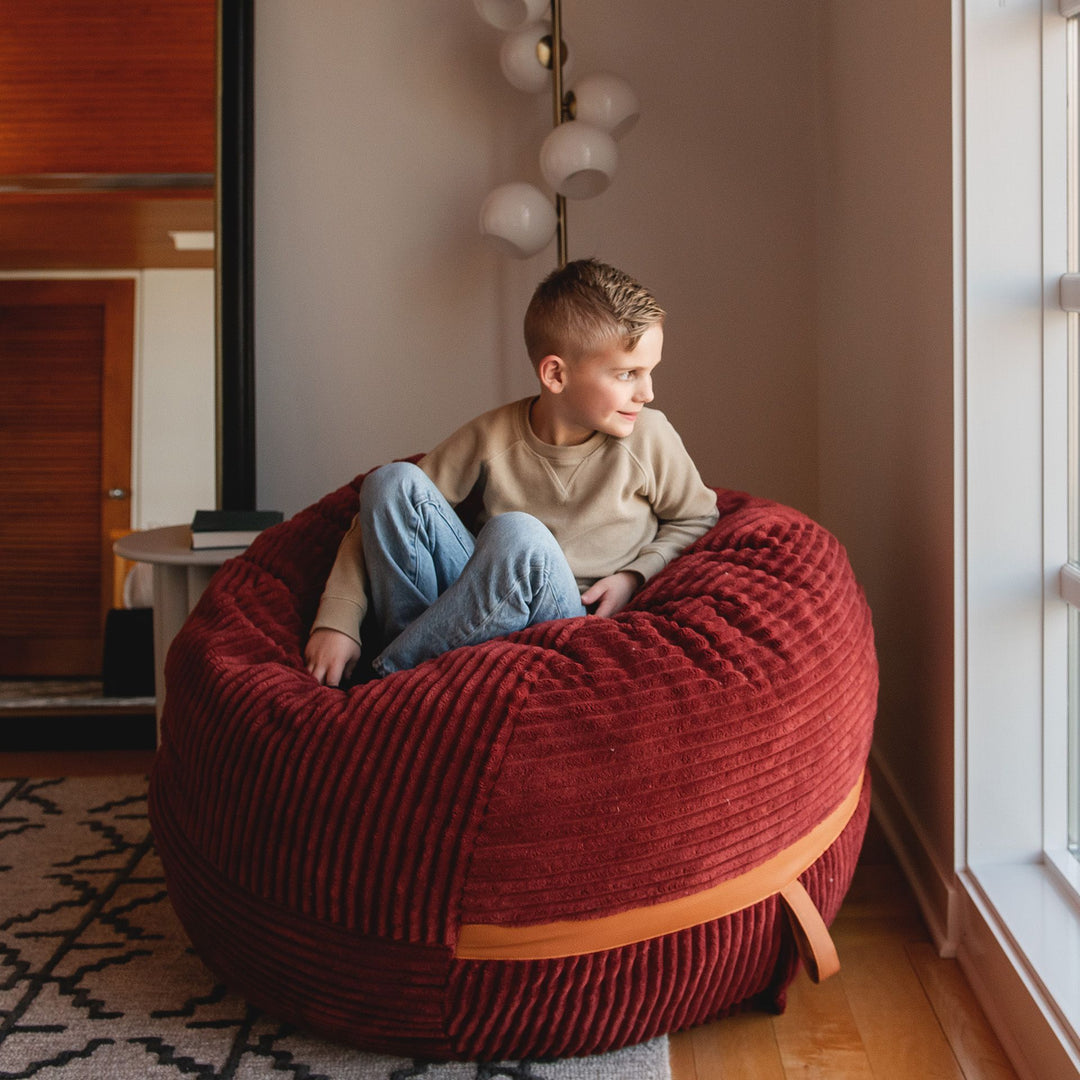 Kid looking out window on medium bean bag chair filled with foam #color_deep-terracotta-corded-plush