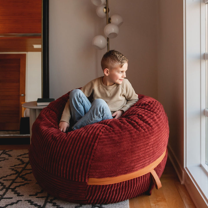 Kid looking out window on medium bean bag chair filled with foam #color_deep-terracotta-corded-plush