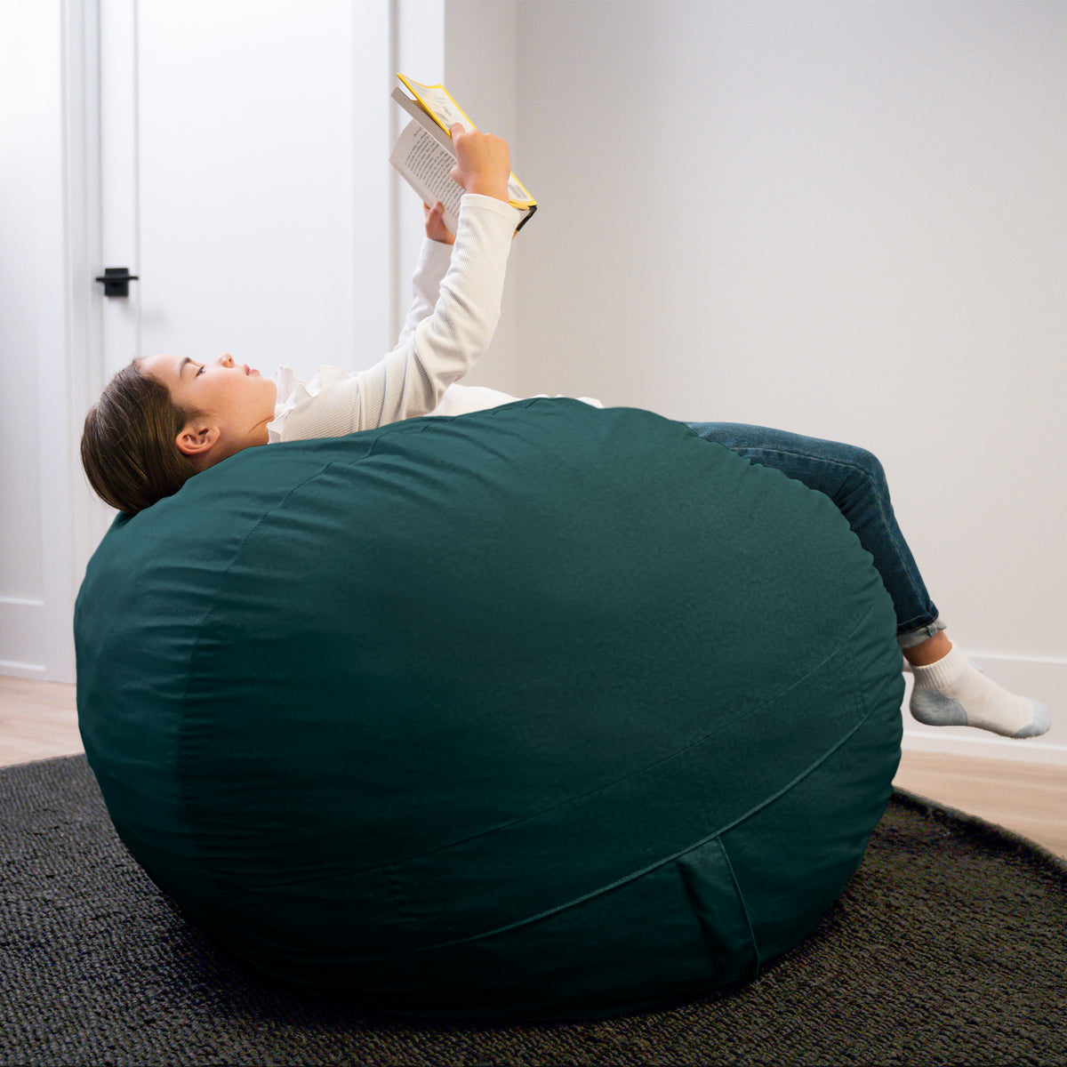 Person lying on a large teal bean bag reading a book in a room with white walls.