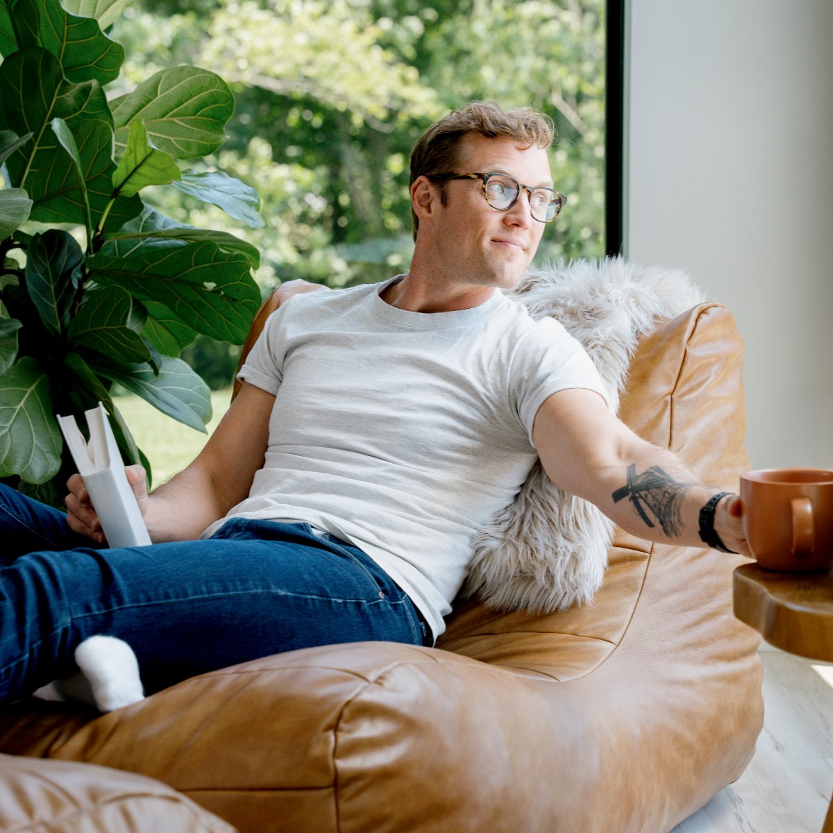 Man sitting on a brown leather chair holding a book and a mug, with a plant and window in the background.