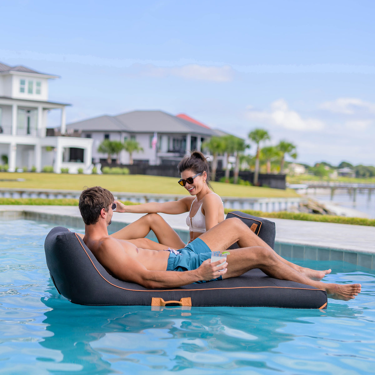 Two people relaxing on a floating lounge in a pool with a scenic background.