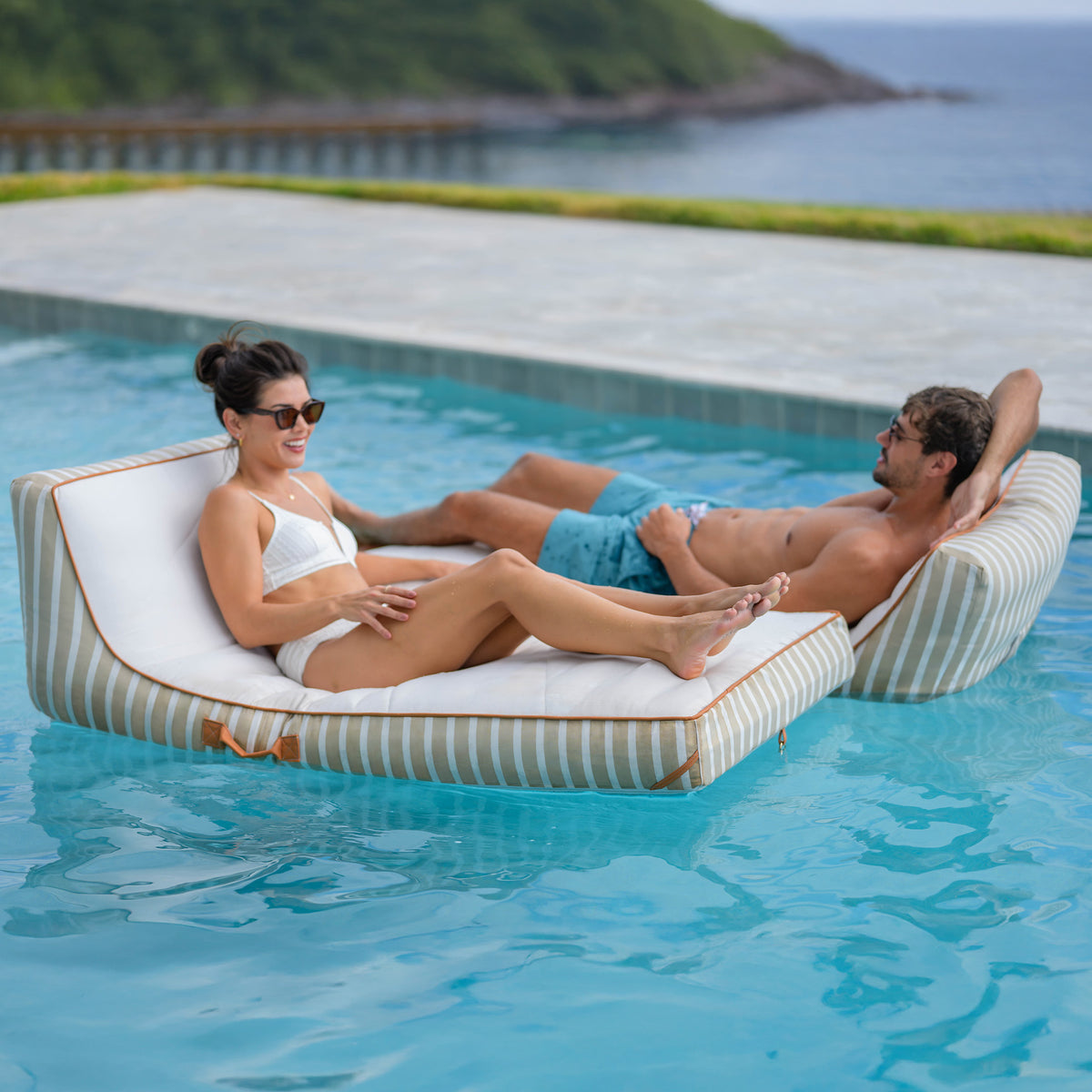 Two people relaxing on a striped pool float in a pool with a scenic background.