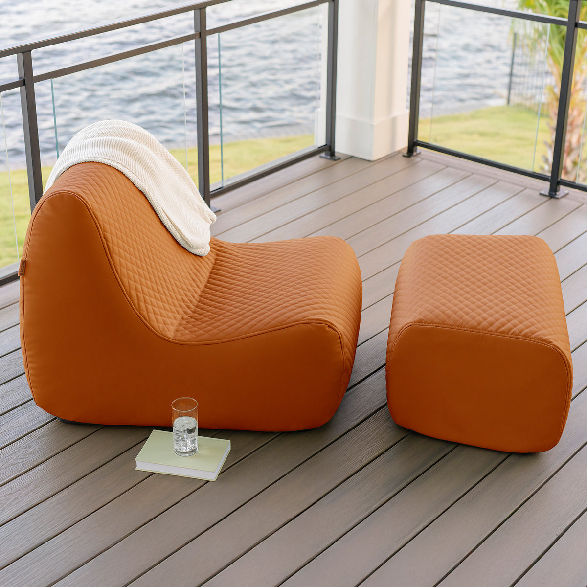 Orange lounge chair and ottoman on a wooden deck with a view of water.