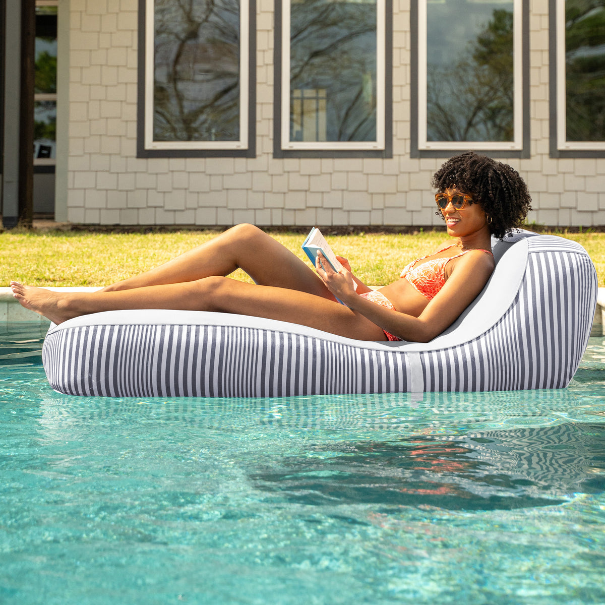 Woman lounging on a striped inflatable pool float in a pool with a house in the background #color_shoreline-gray-stripe-olefin