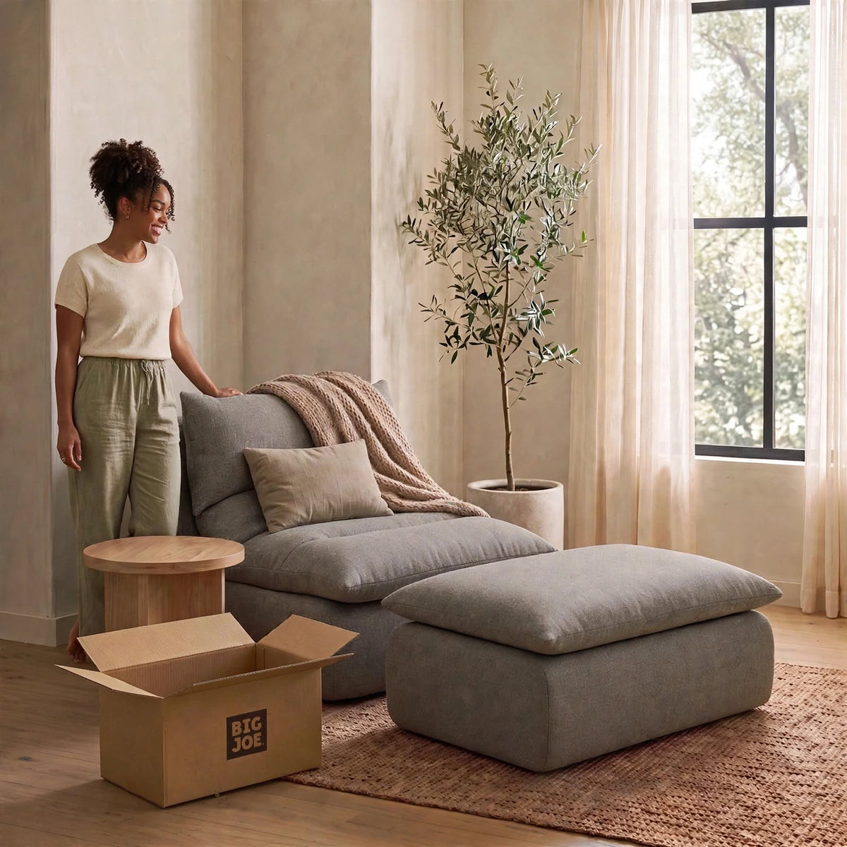 A woman stands beside a large gray upholstered lounge chair with matching ottoman in a softly lit room. A beige throw blanket is draped over the chair, with a potted tree, curtains, window, and an open cardboard moving box visible. #color_misty-gray-terra-knit