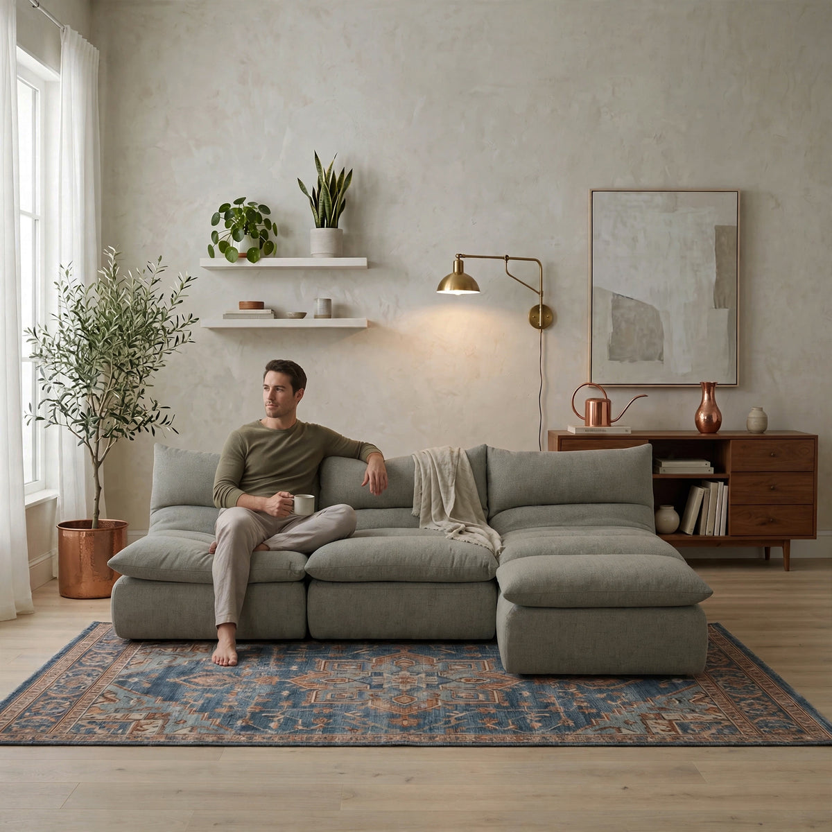A man sits on a large light gray sectional sofa in a modern, well-lit living room. The room features a textured beige wall, a blue patterned rug, a wooden sideboard, and a brass wall lamp. #color_misty-gray-terra-knit