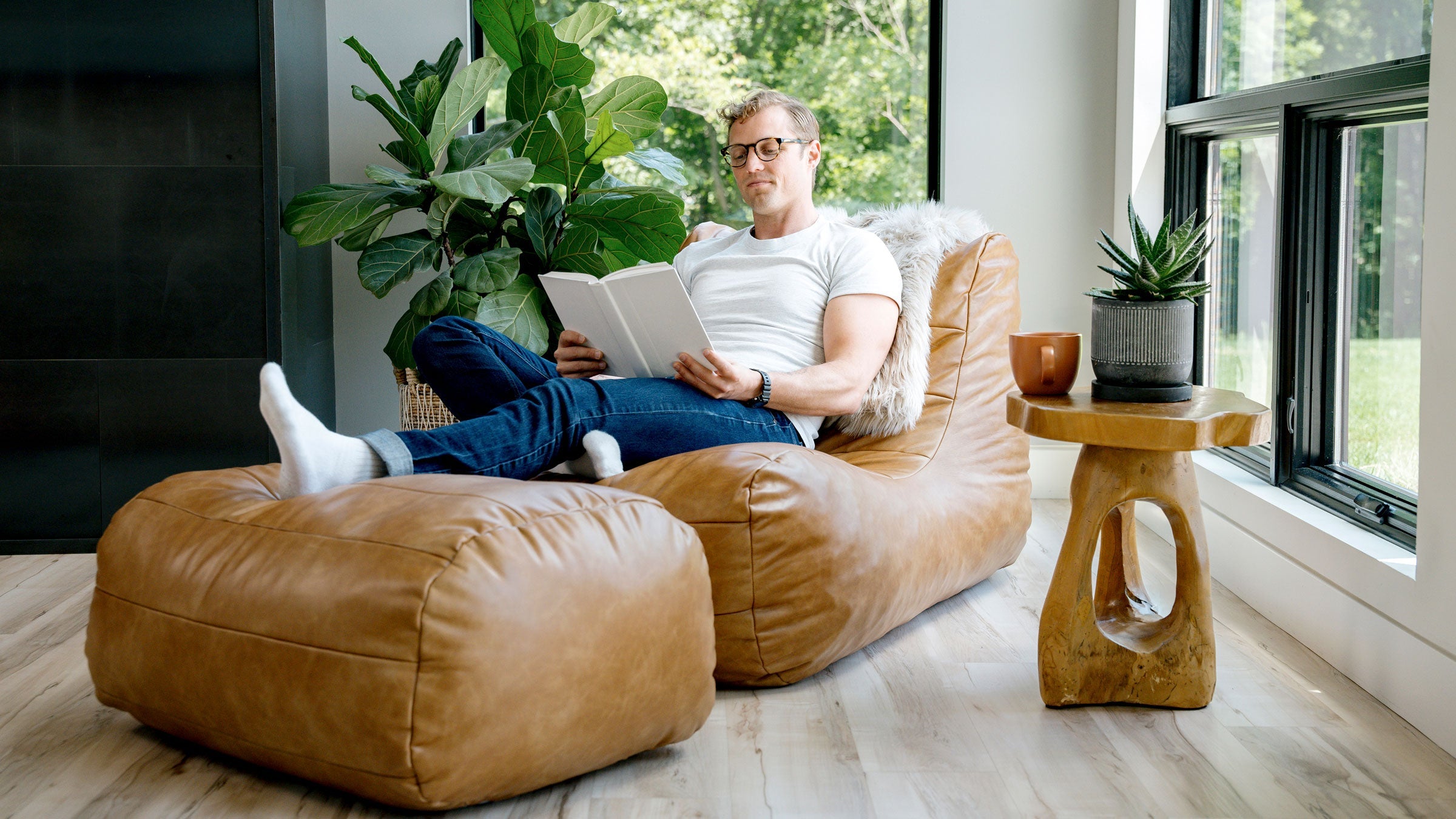 Man relaxing on a lounge chair and ottoman reading a book with window scape and plants in background.