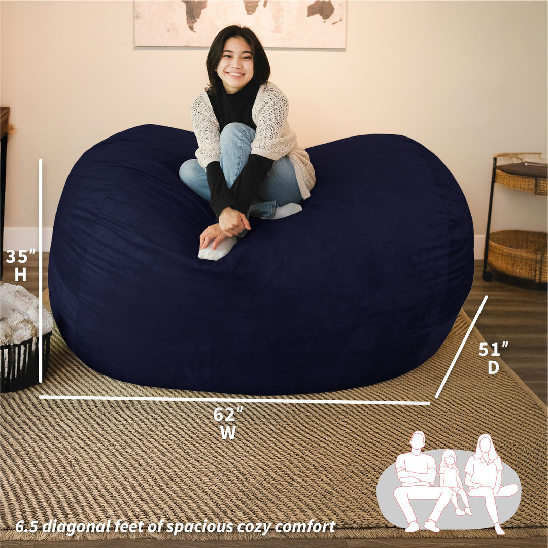 Woman sitting on a large navy bean bag chair with dimensions displayed. #color_midnight-plush