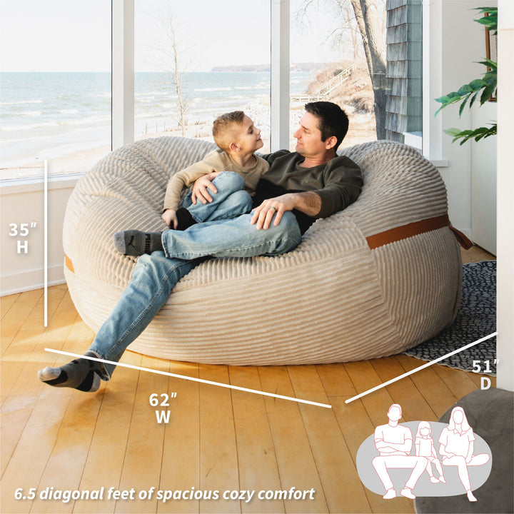 Man and child sitting on a large bean bag chair in a room with windows showing an ocean view. #color_vanilla-latte-corded-plush