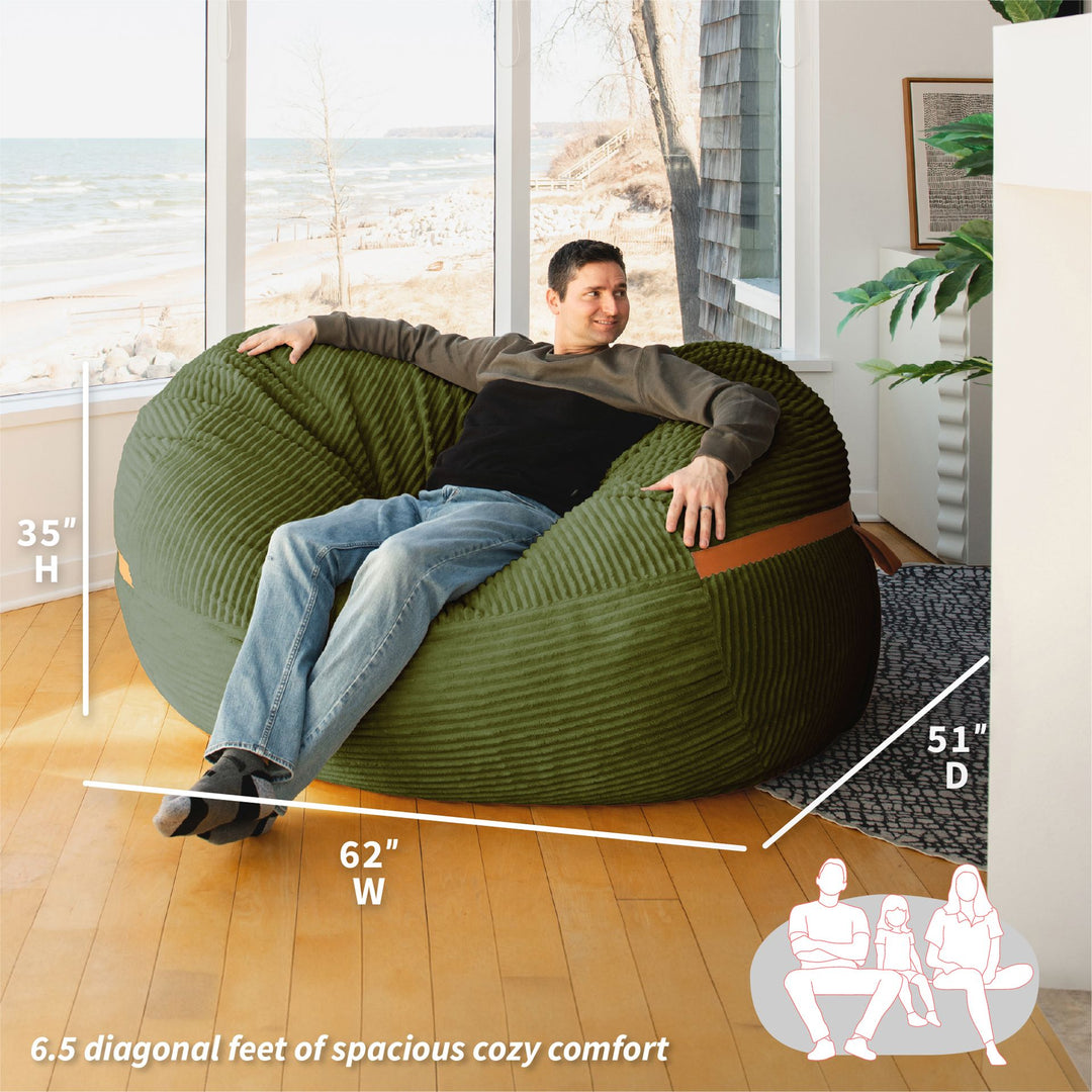 Man sitting in a large green bean bag chair with measurements and illustrations in a room with large windows. #color_moss-green-corded-plush