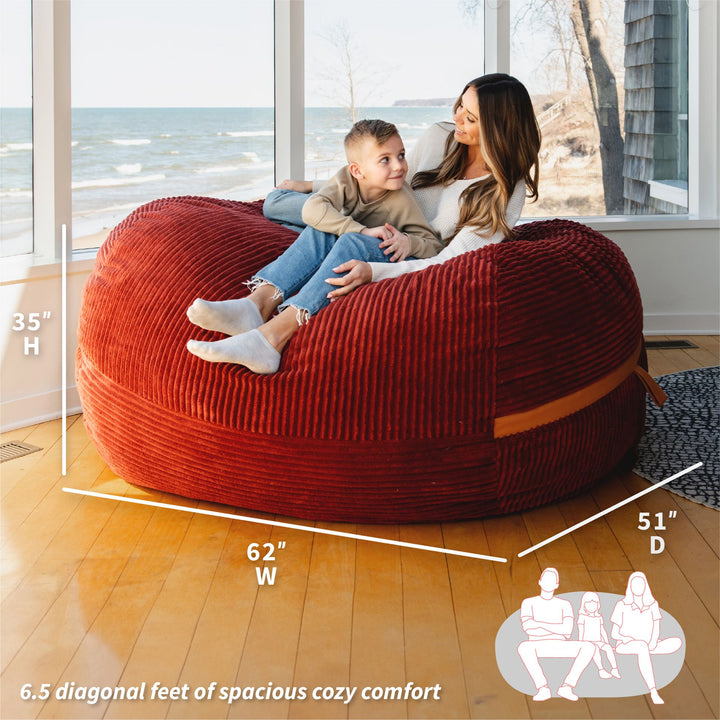 Red bean bag chair with measurements and people sitting on it, near a window with a view of water. #color_deep-terracotta-corded-plush