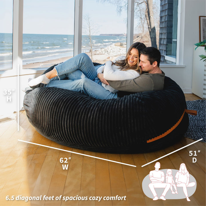 Two people sitting on a large black bean bag chair with measurements and a scenic background. #color_black-licorice-corded-plush