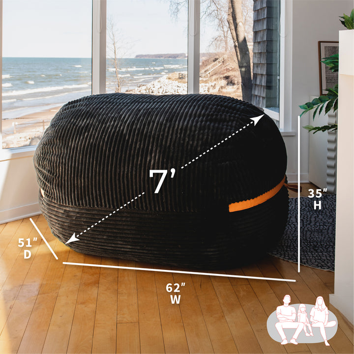 Black bean bag with measurements in a room with large windows showing a beach view. #color_black-licorice-corded-plush