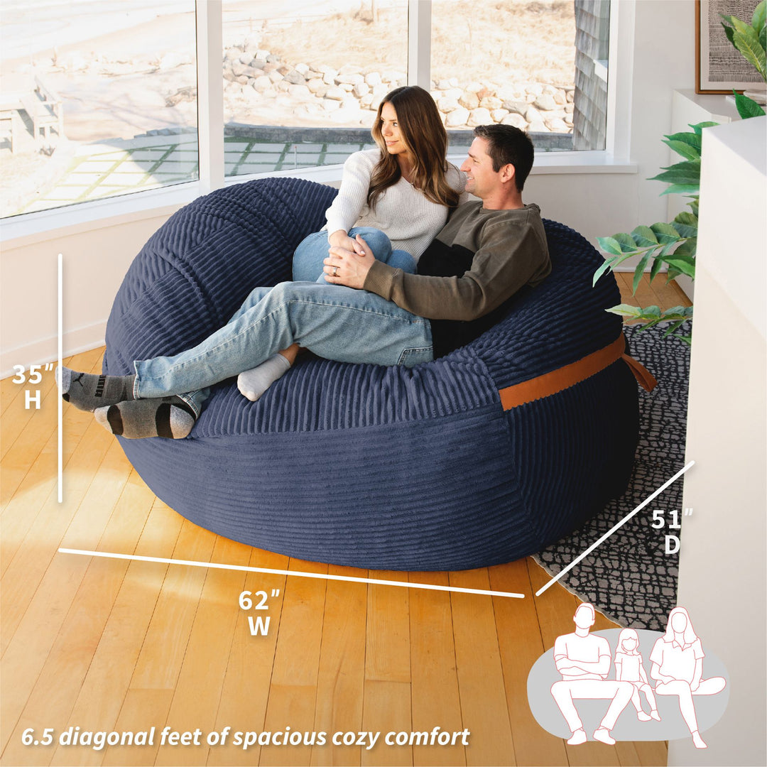 Two people sitting on a large blue bean bag chair with dimensions displayed. #color_midnight-blue-corded-plush