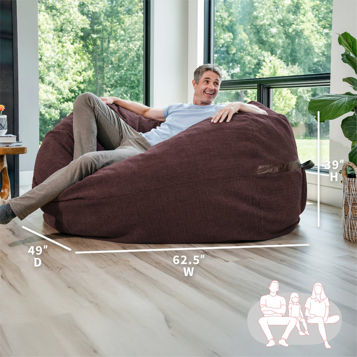 Man sitting on a large brown bean bag chair in a room with large windows. #color_merlot-cloudweave 