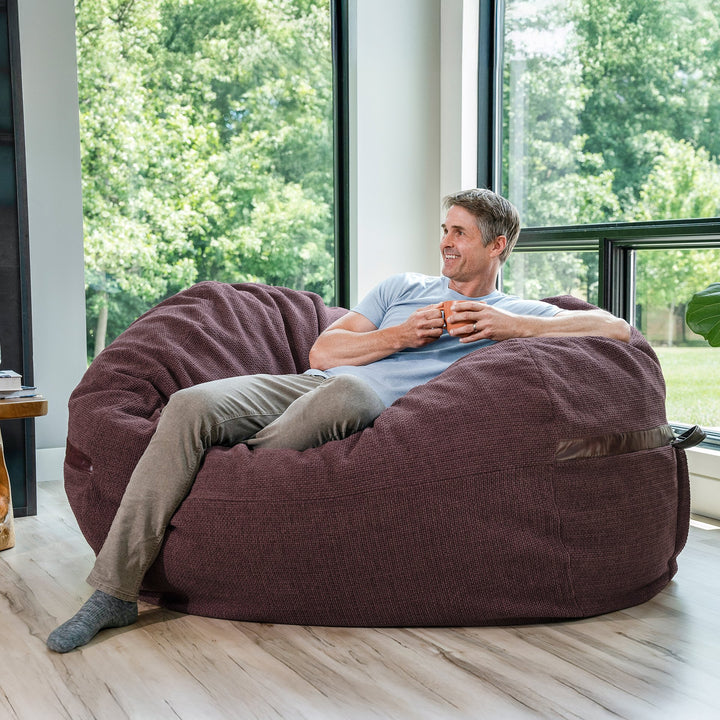 Man sitting on a large maroon bean bag chair in a room with large windows and greenery outside. #color_merlot-cloudweave