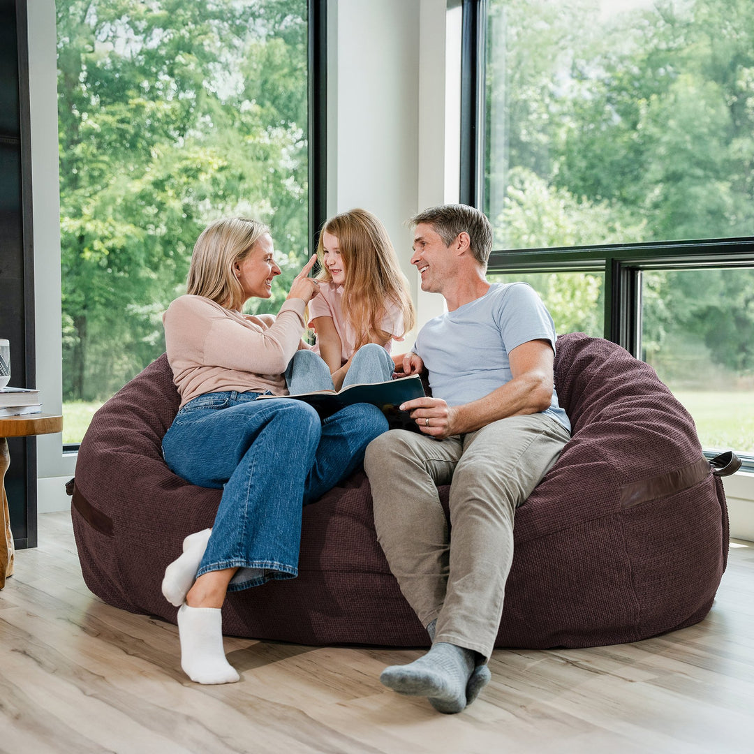 Family sitting on a large maroon bean bag chair in a room with large windows. #color_merlot-cloudweave