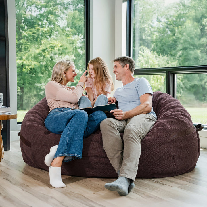 Family sitting on a large maroon bean bag chair in a room with large windows. #color_merlot-cloudweave