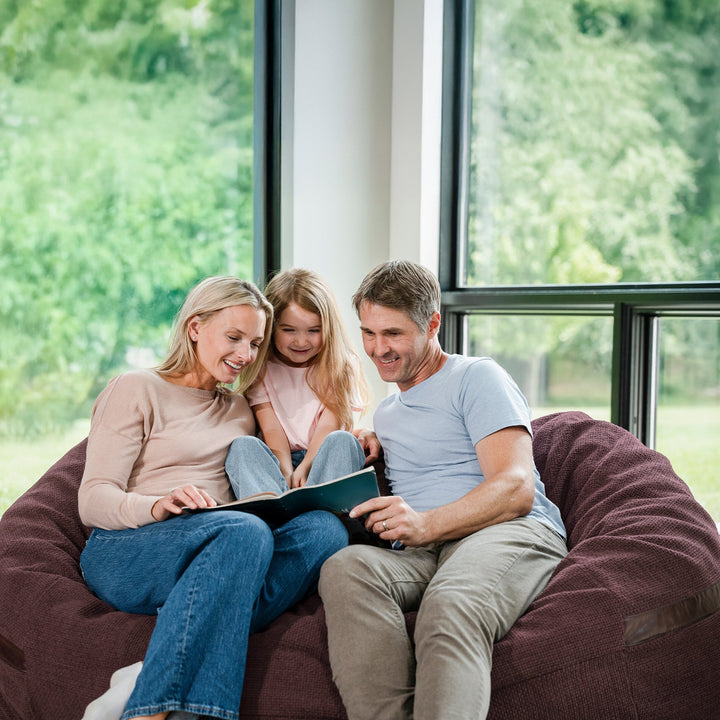 Family of three sitting on a bean bag chair indoors with a view of greenery outside. #color_merlot-cloudweave
