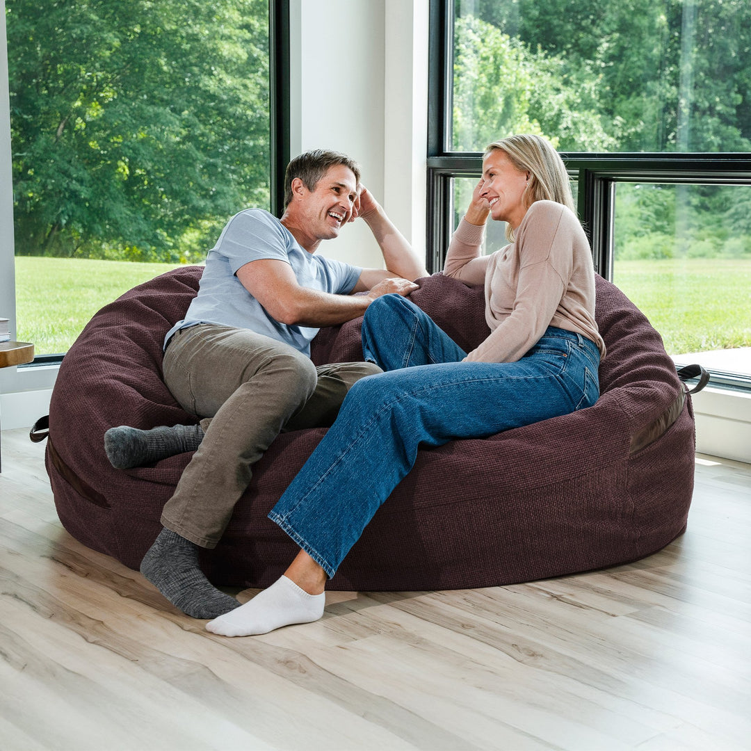 Man and woman sitting on a large brown bean bag chair in a room with large windows. #color_merlot-cloudweave