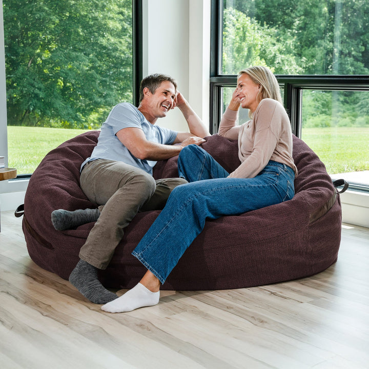 Man and woman sitting on a large brown bean bag chair in a room with large windows. #color_merlot-cloudweave