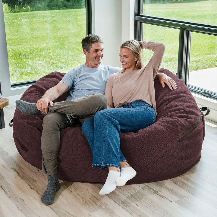 Man and woman sitting on a large bean bag chair in a room with large windows. #color_merlot-cloudweave