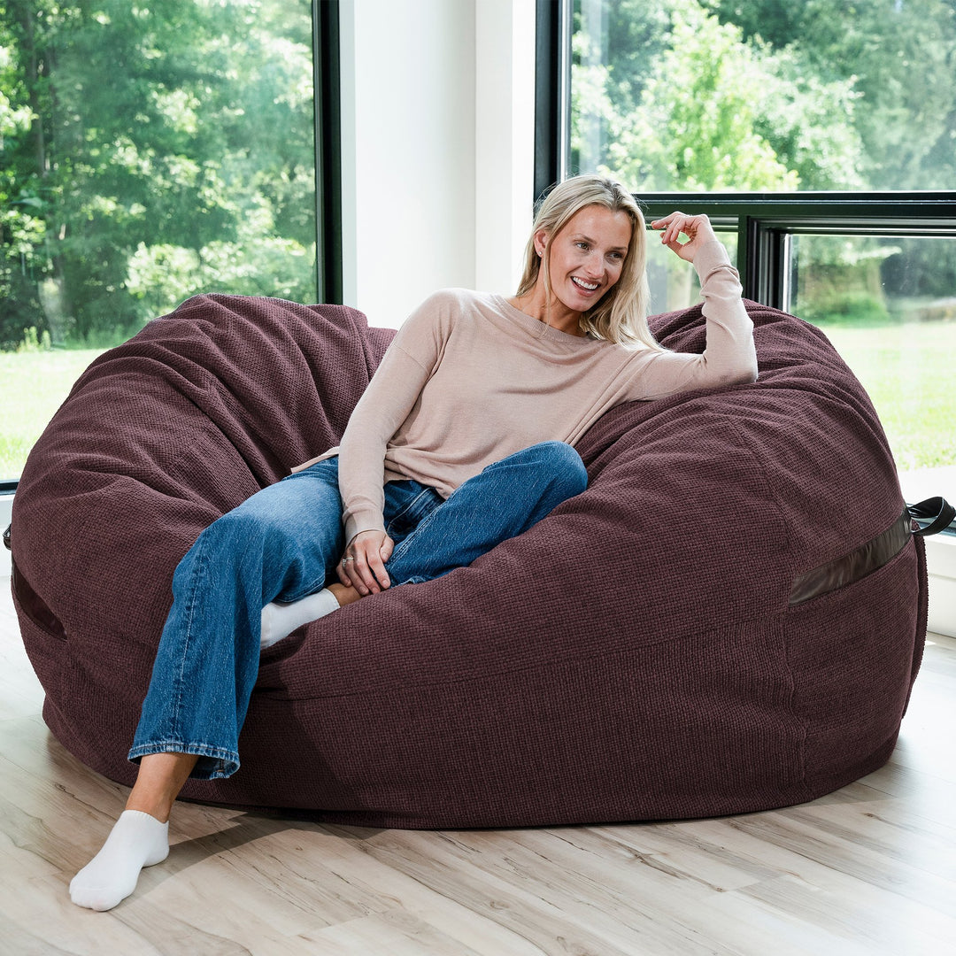 Woman sitting on a large maroon bean bag chair in a room with large windows. #color_merlot-cloudweave