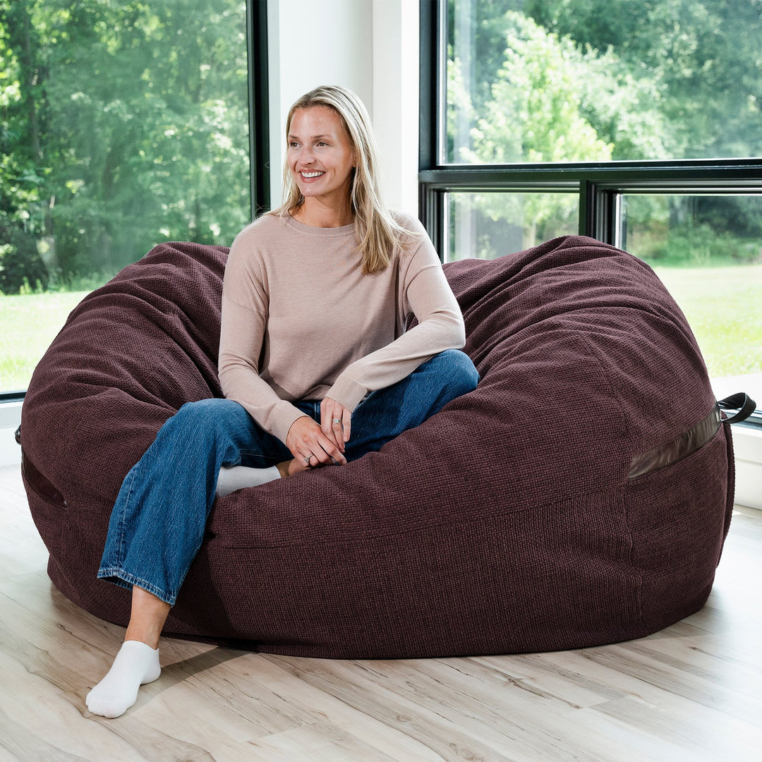 Woman sitting on a maroon brown bean bag chair in a room with large windows. #color_merlot-cloudweave