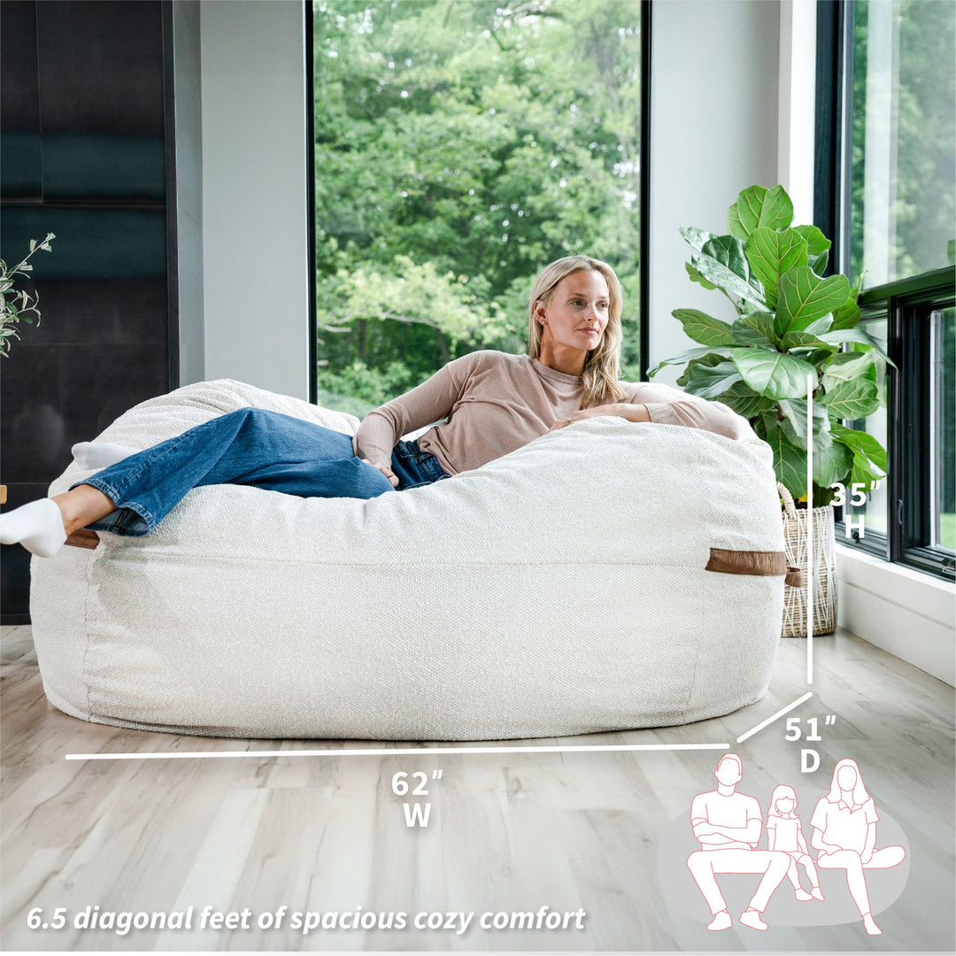 Woman relaxing on a large bean bag chair with dimensions displayed #color_sandstone-textured-boucle