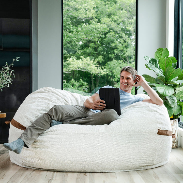 Man relaxing on a bean bag chair indoors with a tablet. #color_sandstone-textured-boucle