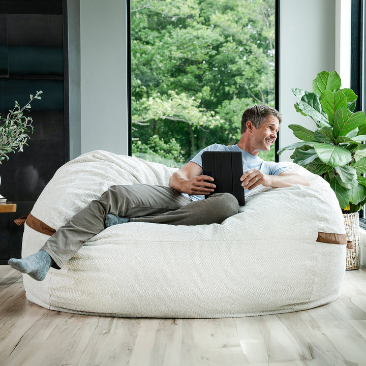 Man sitting on a large white bean bag chair indoors, holding a tablet. #color_sandstone-textured-boucle