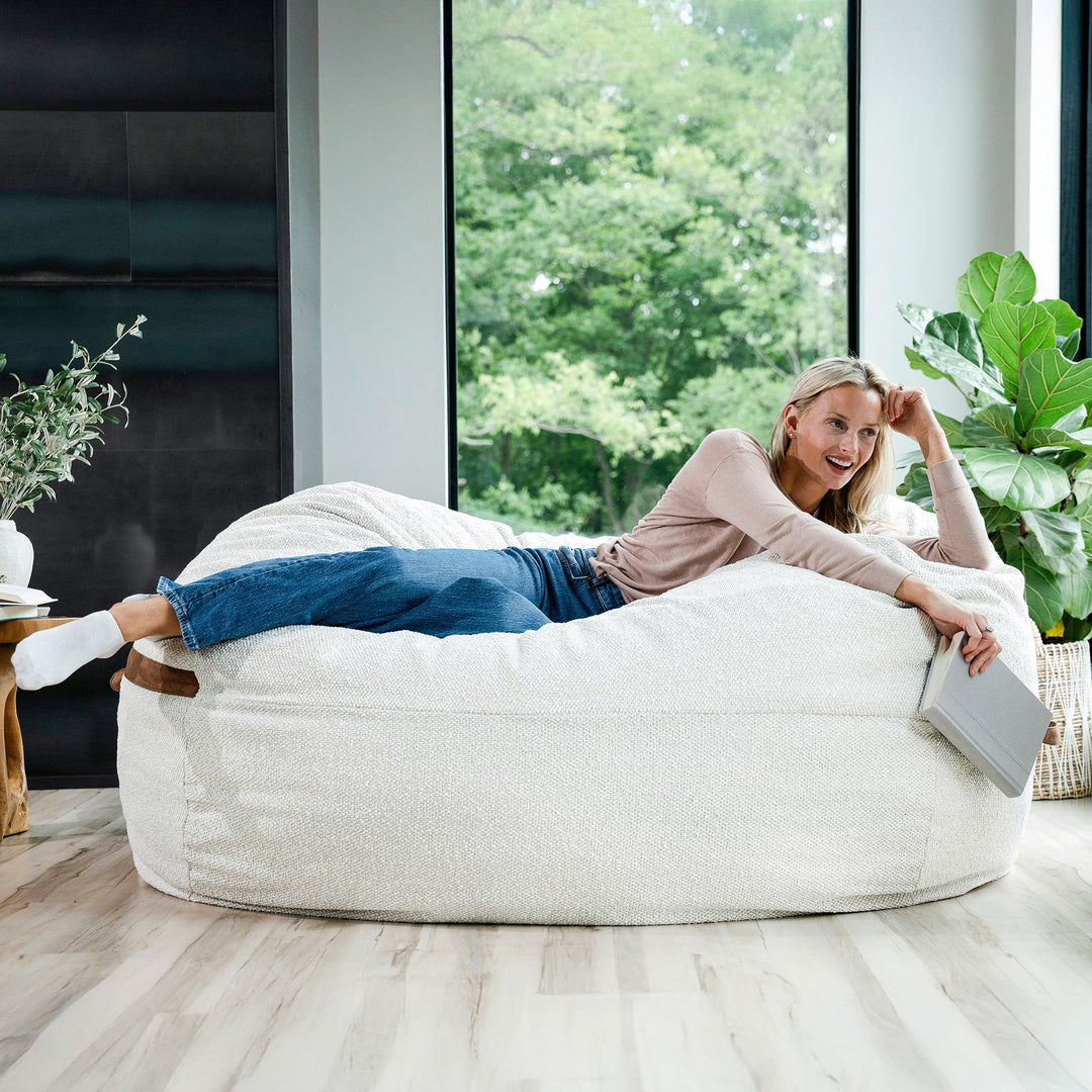 Woman lying on a large bean bag chair in a modern living room with large windows. #color_sandstone-textured-boucle