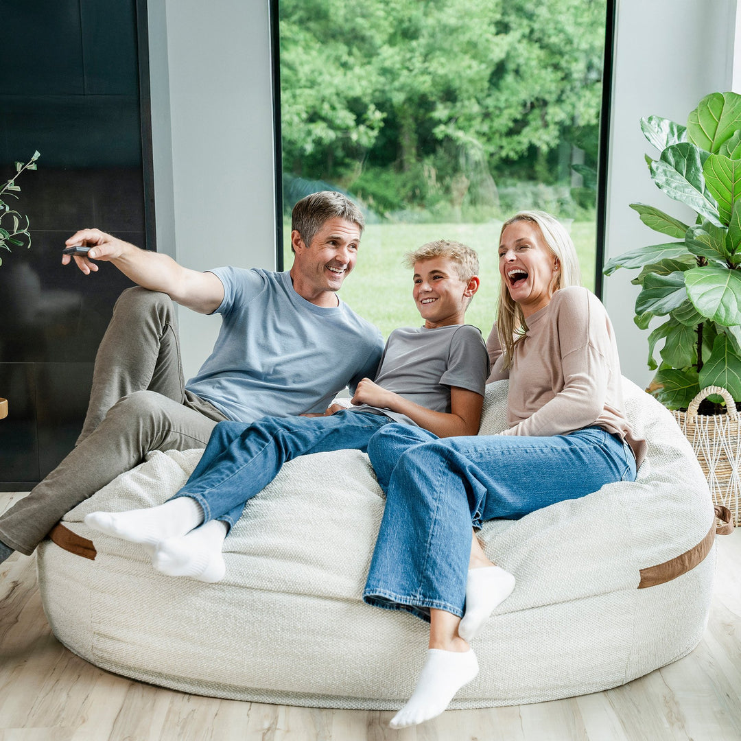 Family of three sitting on a large bean bag in a modern living room. #color_sandstone-textured-boucle