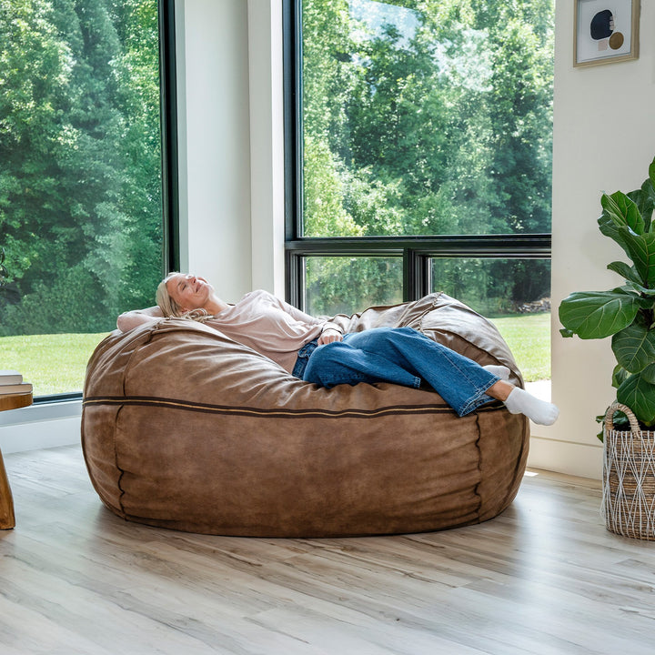 Person relaxing on a large brown bean bag chair in a room with large windows and greenery outside. #color_coffee-soft-vegan-leather