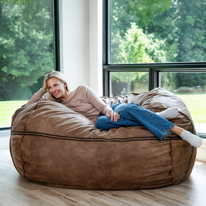 Woman sitting on a large brown bean bag chair in a room with large windows. #color_coffee-soft-vegan-leather