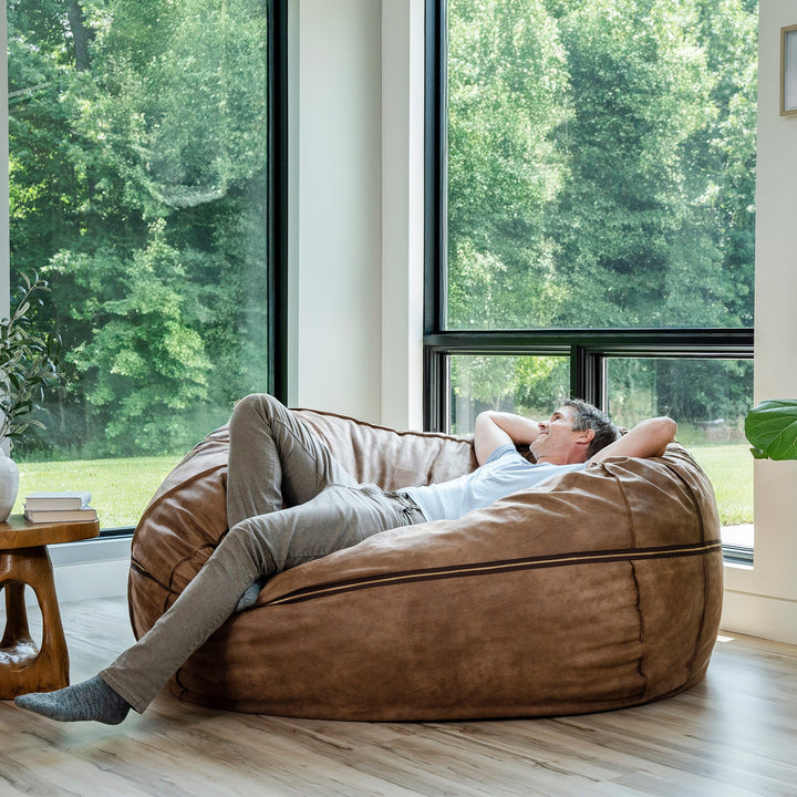 Man relaxing on a large brown bean bag chair in a room with large windows and greenery outside. #color_coffee-soft-vegan-leather
