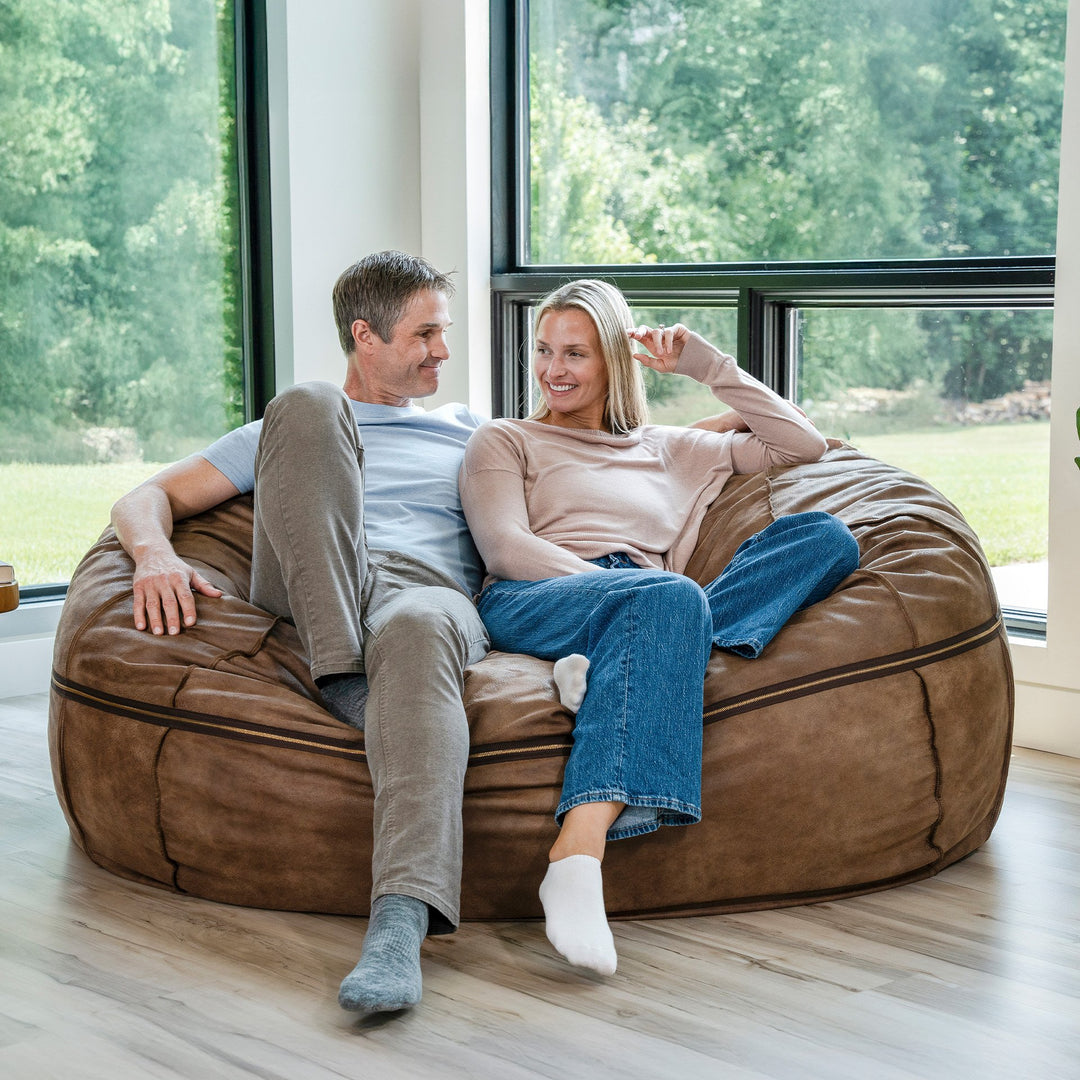 Man and woman sitting on a large brown bean bag chair in a room with large windows. #color_coffee-soft-vegan-leather