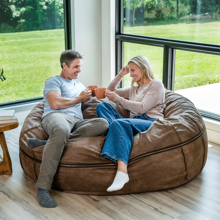 Man and woman sitting on a large brown bean bag chair indoors with a view of greenery outside. #color_coffee-soft-vegan-leather