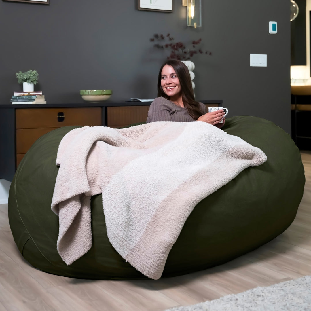 Woman sitting on a large green bean bag with a blanket, holding a mug in a room. #color_forest-green-velveteen
