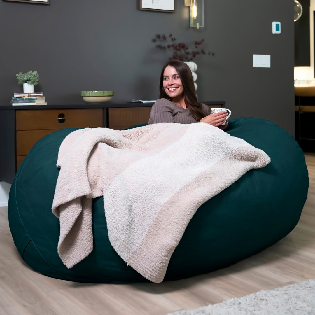 Woman sitting on a large green bean bag with a blanket, holding a mug in a room. #color_deep-sea-velveteen