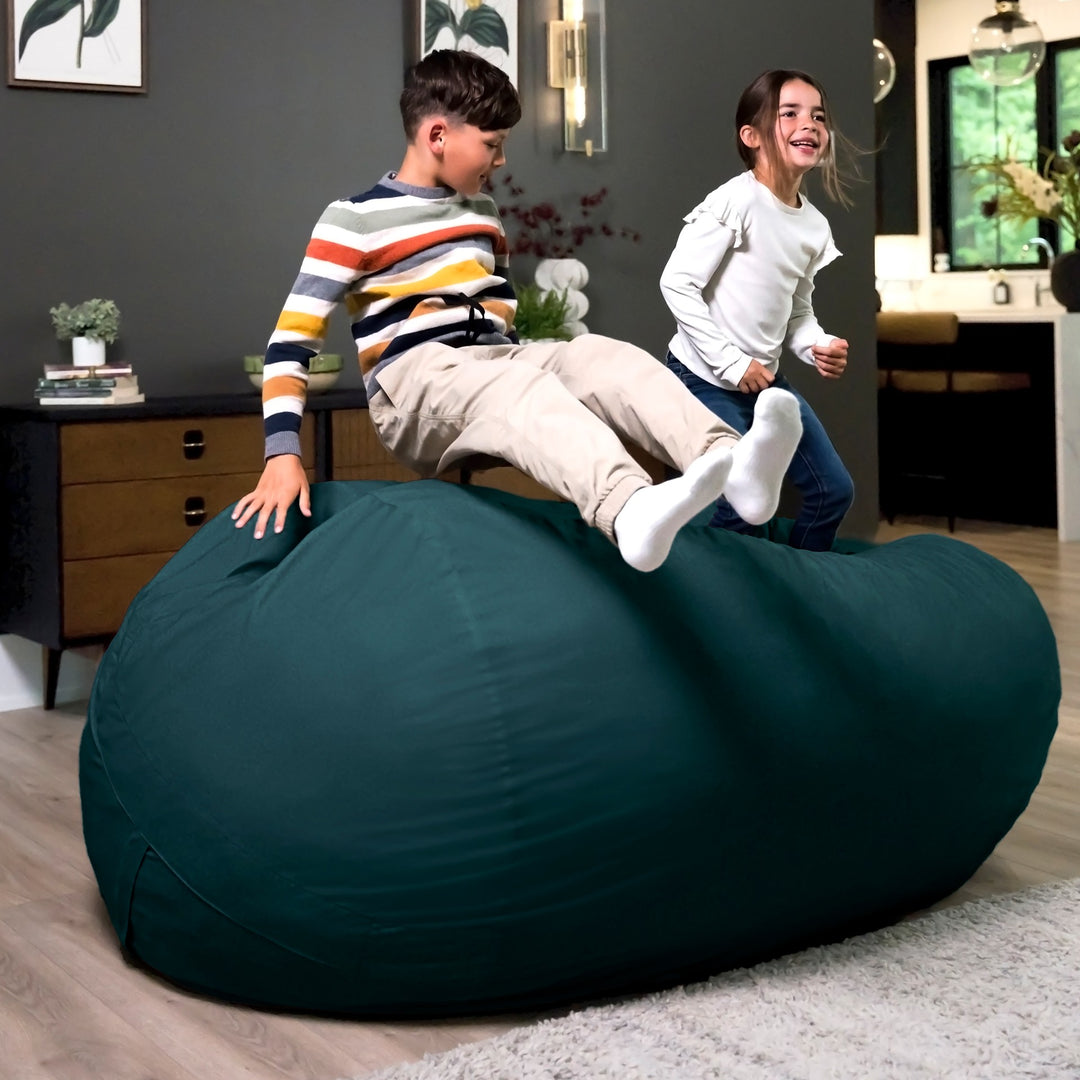 Two children playing on a large teal bean bag in a room with a desk and plants. #color_deep-sea-velveteen