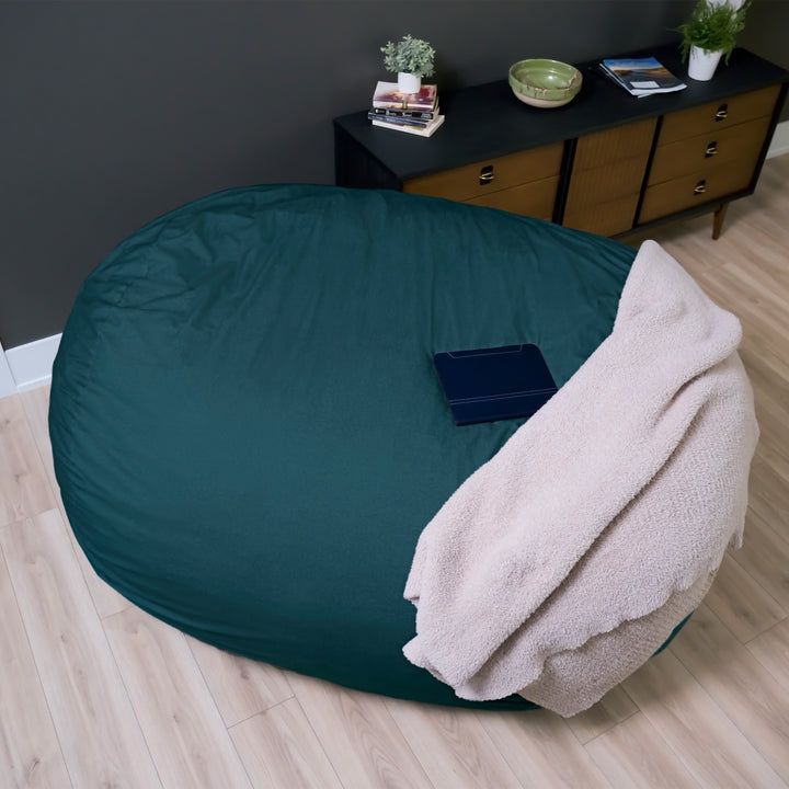 Large green bean bag chair with a beige blanket on a wooden floor in a room with a dark wall and a dresser. #color_deep-sea-velveteen
