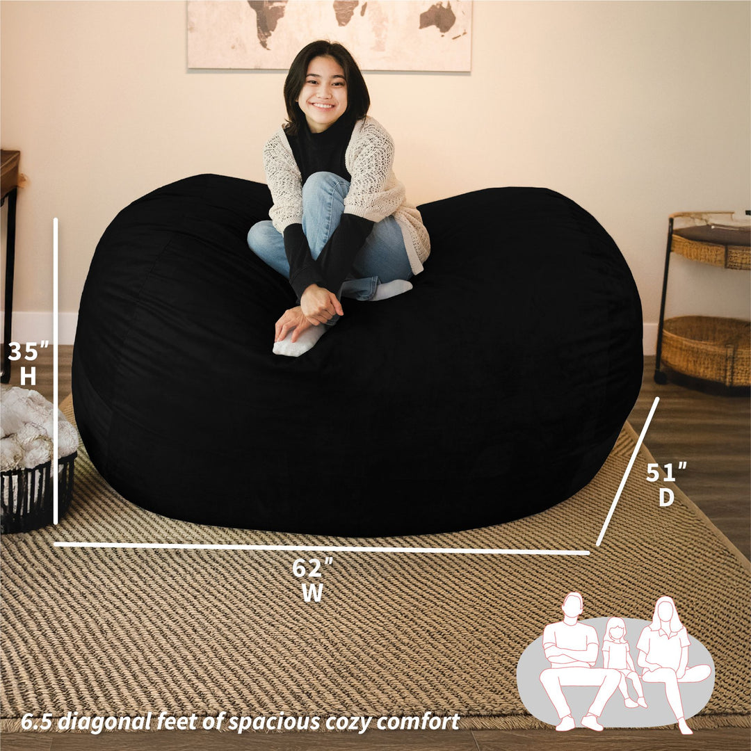 Woman sitting on a large black bean bag chair with measurements and illustrations in a living room setting. #color_black-plush