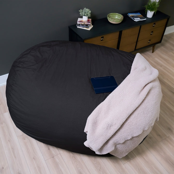 Black bean bag chair with a beige blanket on a wooden floor, with a dark wall and wooden cabinet in the background. #color_black-brushed-denim