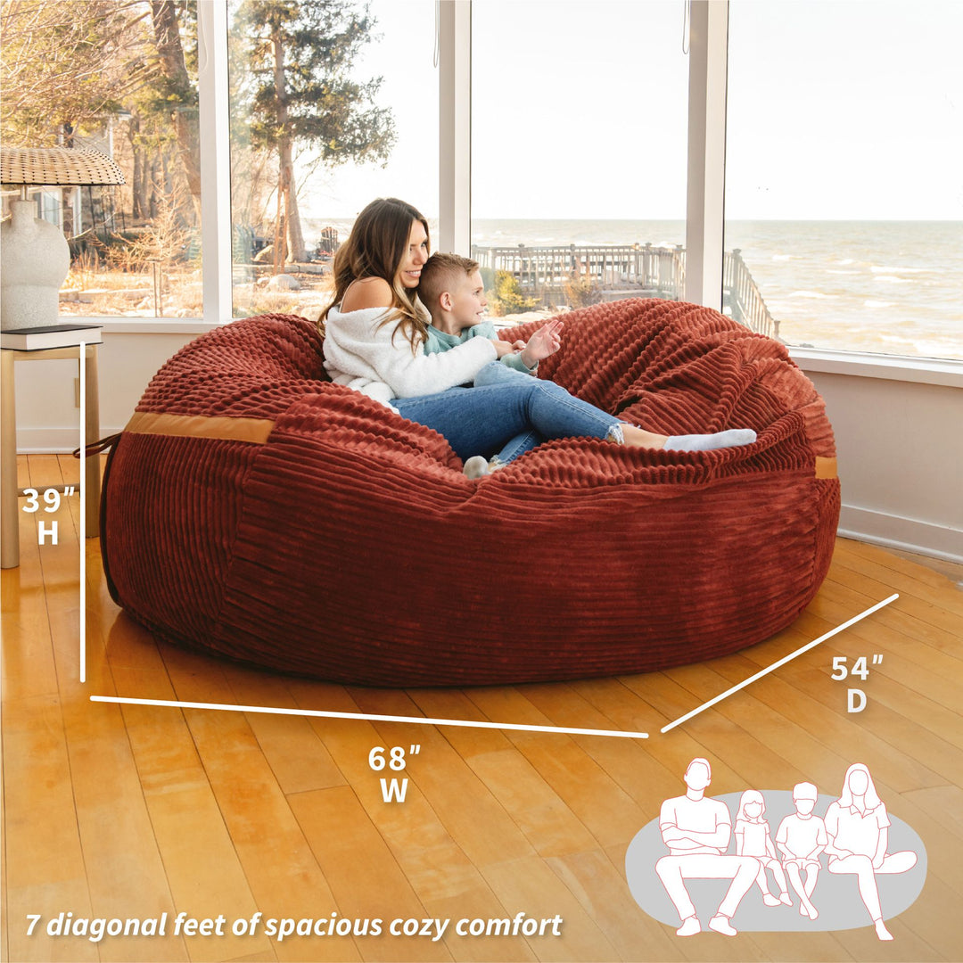Large red bean bag chair with dimensions and people sitting on it in a room. #color_deep-terracotta-corded-plush